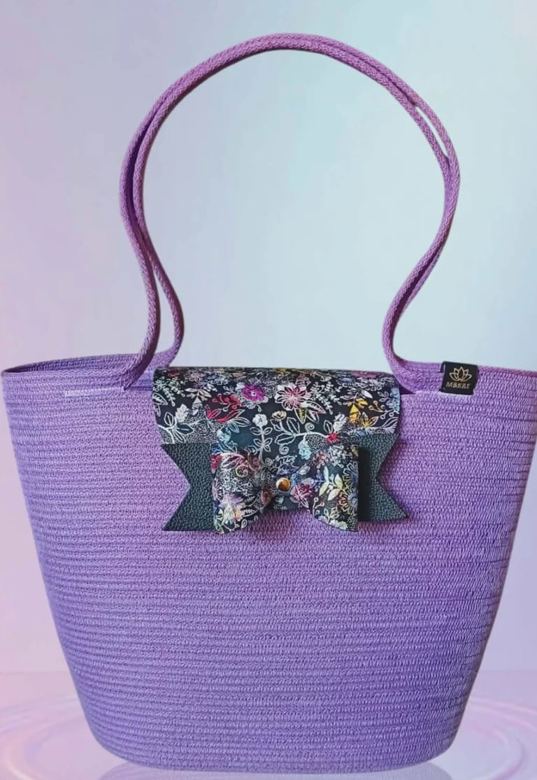 Purple woven tote bag with long handles and a decorative black floral bow accent on the front