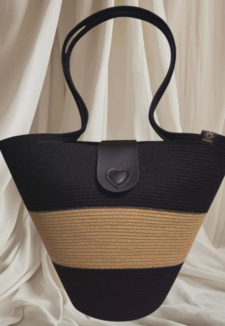Black and tan striped straw tote bag with heart-shaped clasp, displayed against white curtain backdrop