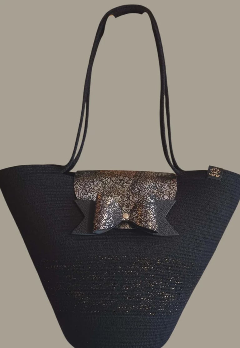 Black woven tote bag with metallic bronze bow accent on beige background